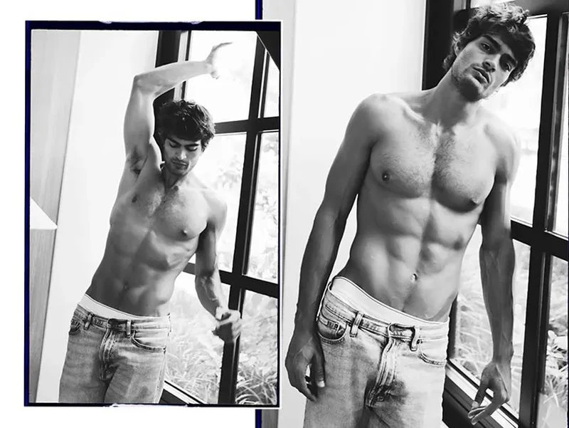 Black and white studio shot of a shirtless adult male model in jeans posing by a window, showcasing a fashion and editorial aesthetic.