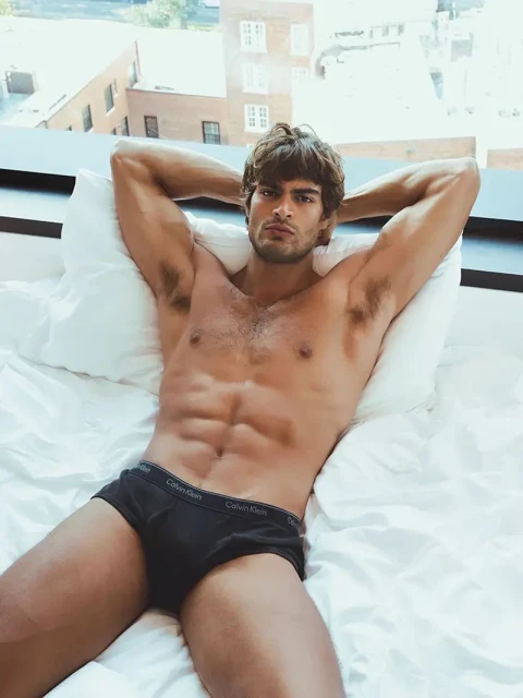 Fit man in underwear relaxing on a white bed, with hands behind his head. Light streams in from a window with a city view.