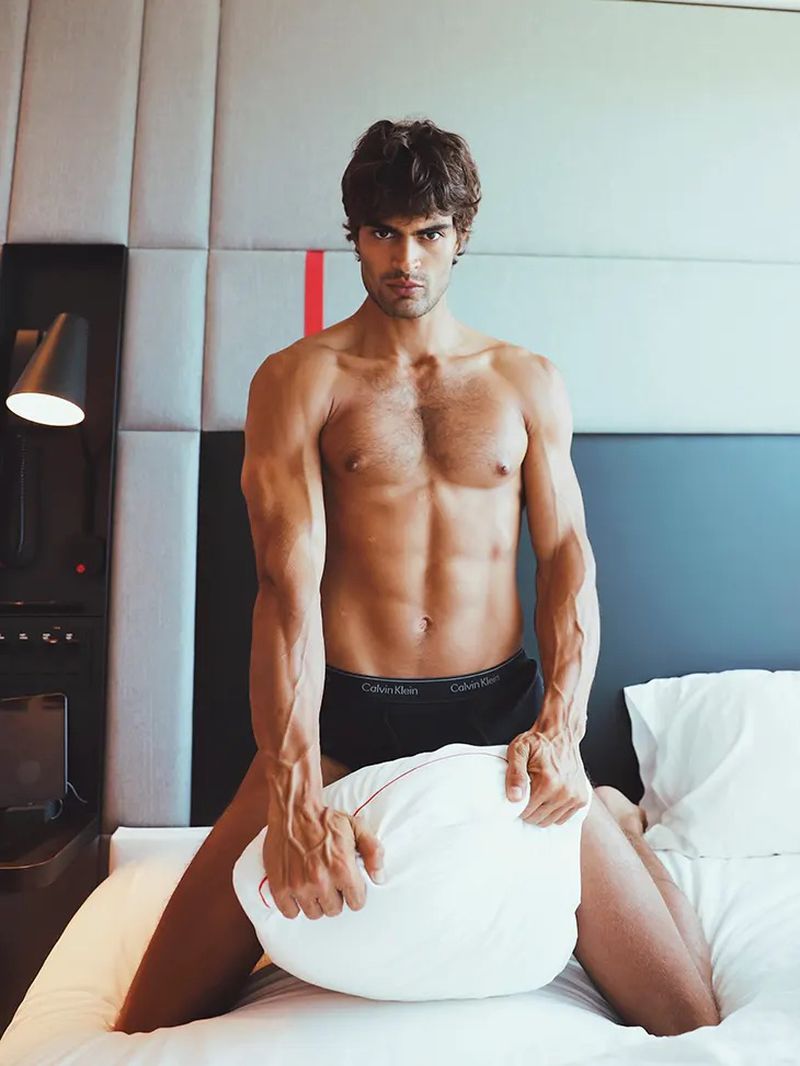 Shirtless man wearing underwear poses kneeling on a white bed, holding a pillow, in a brightly lit indoor setting.