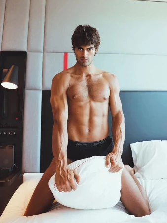Shirtless man wearing underwear poses kneeling on a white bed, holding a pillow, in a brightly lit indoor setting.