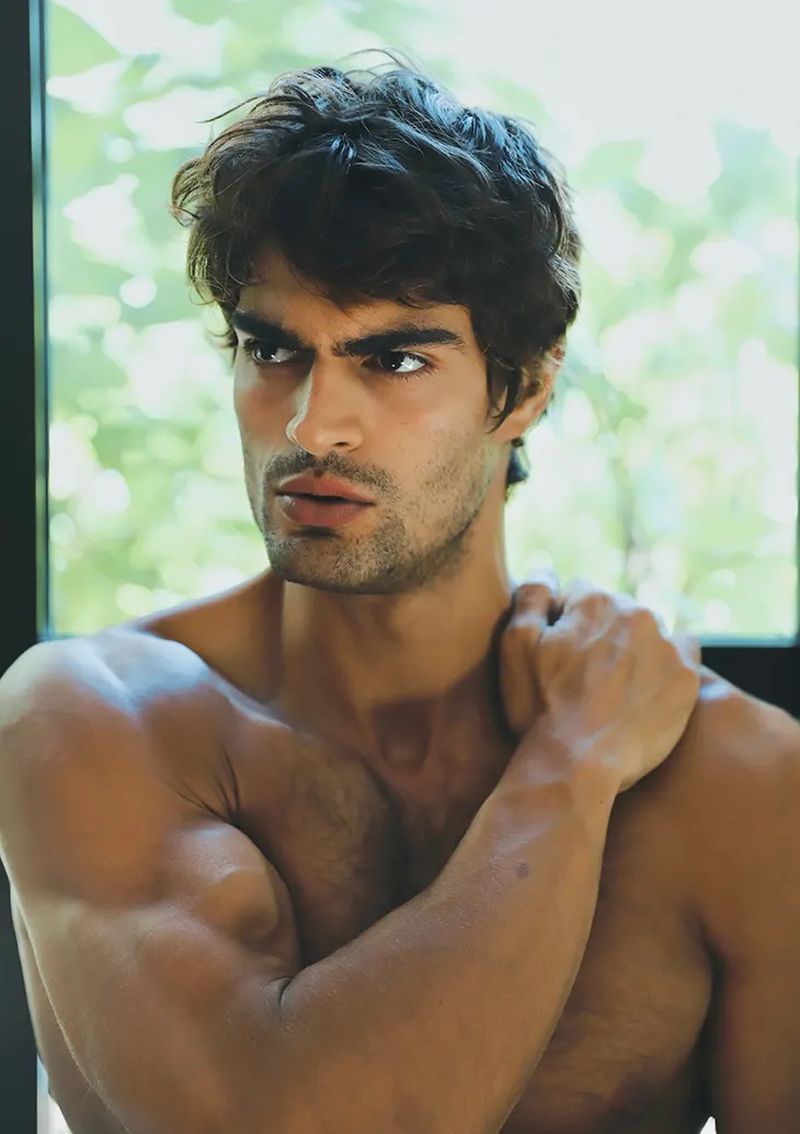 Portrait of a muscular adult male with brown hair, posing shirtless against a blurred green window. Studio lighting and soft shadows.