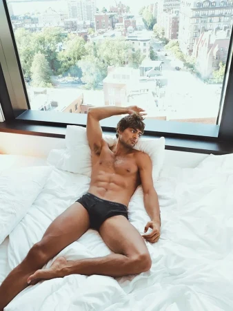 Model Iago Botelho reclining in black briefs on a white bed in front of a large sunlit window overlooking a city street.