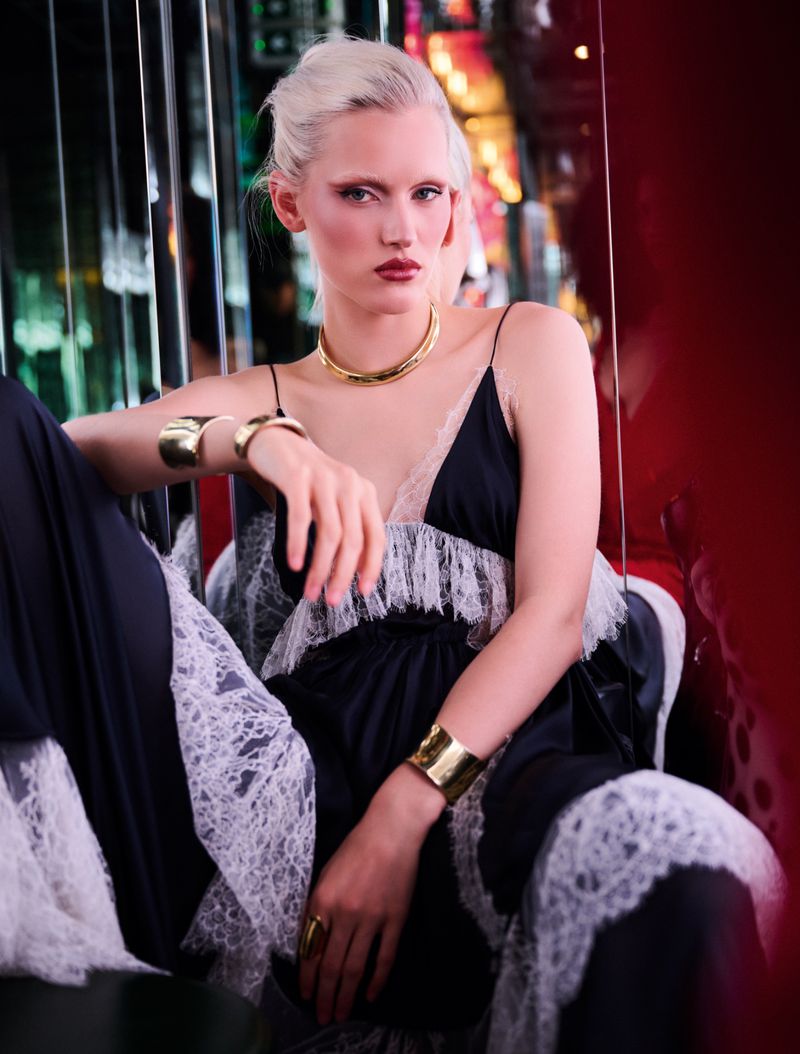 Fashion photo of a platinum blonde woman wearing a black and white lace dress with gold jewellery sitting indoors with red and green lighting.