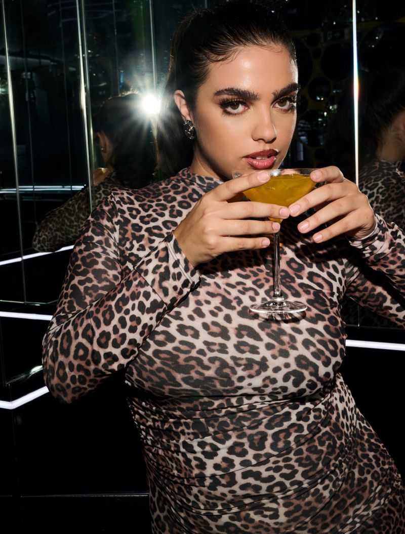 Woman in leopard print dress poses indoors, holding a cocktail in a martini glass with reflective glass and dark lighting.