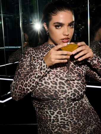 Woman in leopard print dress poses indoors, holding a cocktail in a martini glass with reflective glass and dark lighting.