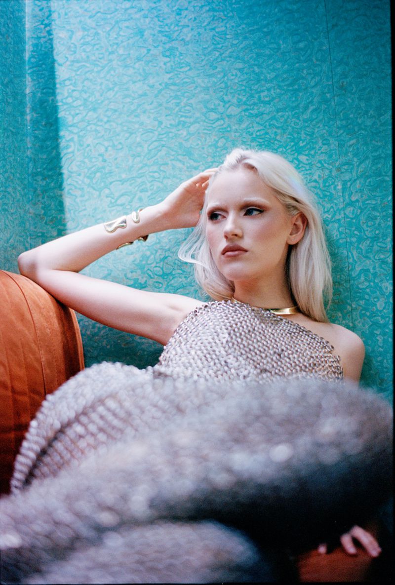 Portrait of a young woman with platinum blonde hair wearing a metallic top and a gold choker, seated on an orange chair.