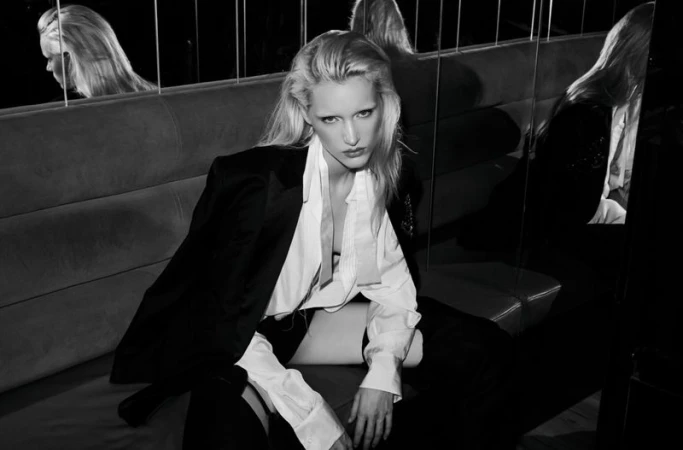 Black and white portrait of a blonde adult woman in a white blouse and dark jacket sitting on a couch in an indoor setting.