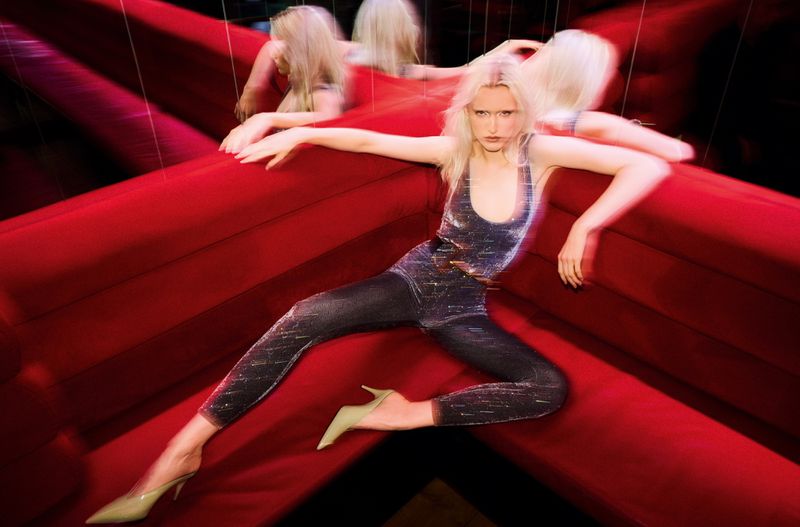 A woman in a shimmering bodysuit poses on a red surface with mirrored reflections, showcasing a dynamic and distorted visual effect.