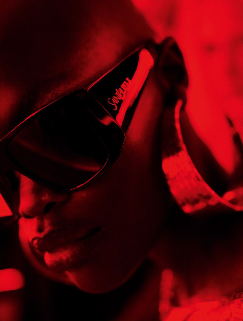 Close-up shot of a woman with dark skin wearing sunglasses and large hoop earrings in vivid red lighting creating a dramatic effect.