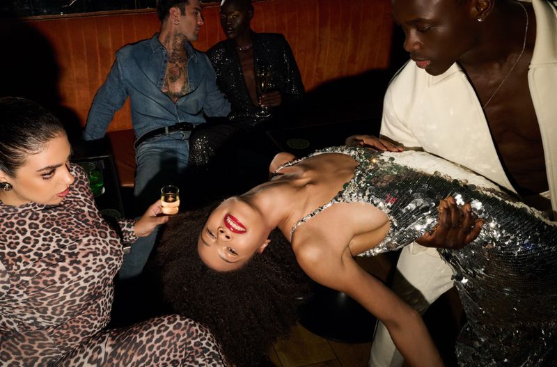 Group of four people in a dark club or lounge, with one woman in a silver dress being held by a man, another woman holding a drink.