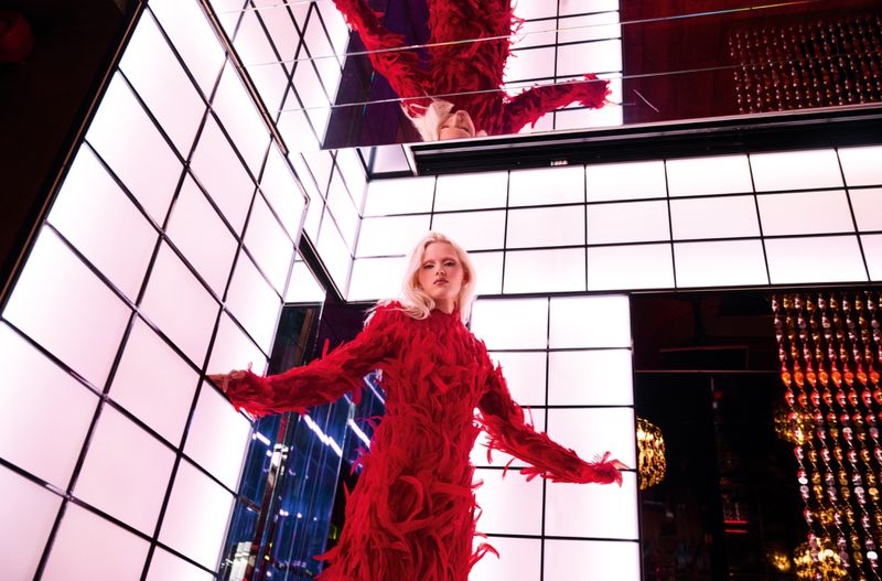 Blonde model poses in a vibrant red feathered dress, surrounded by geometric grid-like lighting in a mirrored photo studio.