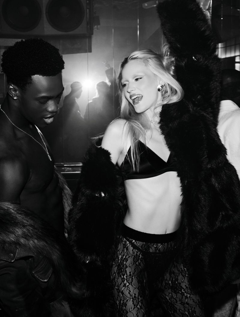 Monochrome fashion portrait of an adult woman wearing a black bra and lace pants with a fur coat standing next to a man at a nightclub.