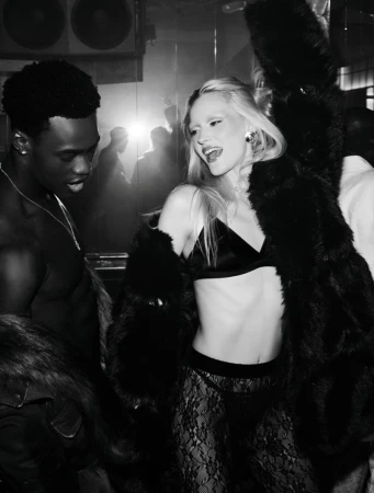 Monochrome fashion portrait of an adult woman wearing a black bra and lace pants with a fur coat standing next to a man at a nightclub.
