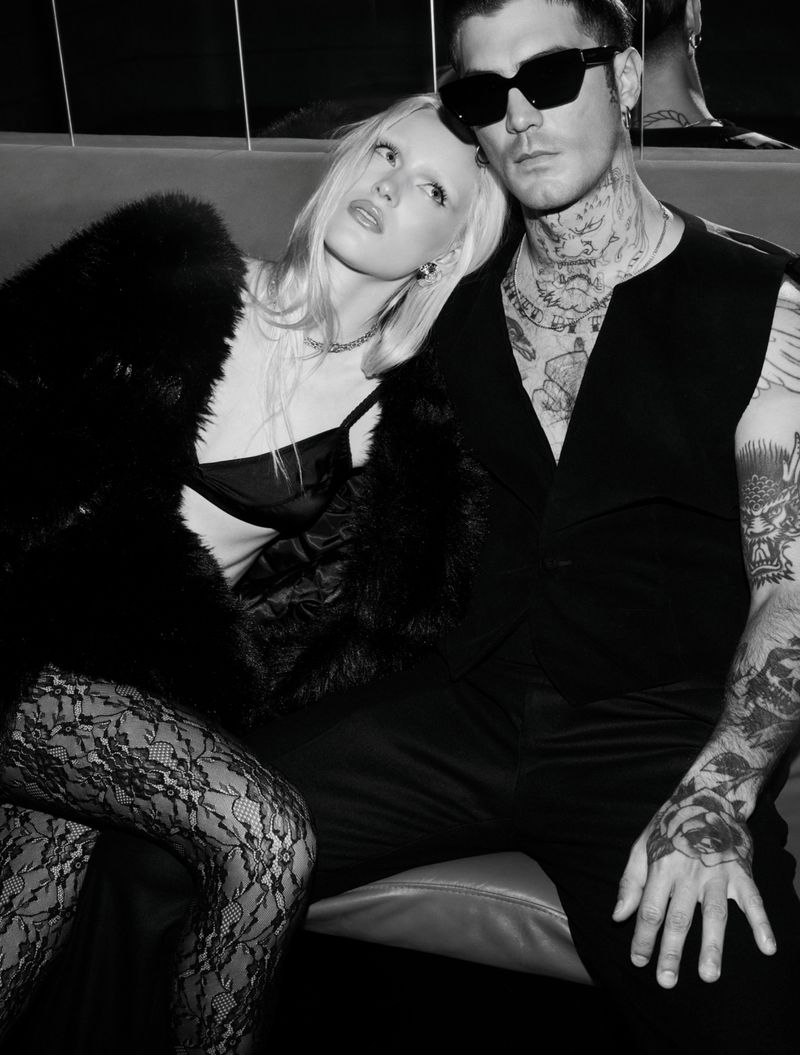 Black and white image of a couple sitting intimately. The tattooed man wears sunglasses, while the woman dons a fur coat and lace tights.