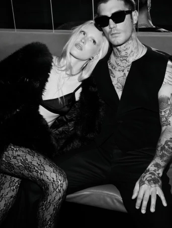 Black and white image of a couple sitting intimately. The tattooed man wears sunglasses, while the woman dons a fur coat and lace tights.
