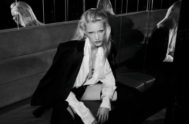 Black and white photo of model Ine Michelmann wearing an open white shirt and black blazer seated in front of panelled mirrors.