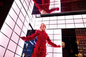 Model Ine Michelmann poses in a textured red feather dress surrounded by illuminated grid walls and ceiling mirrors.