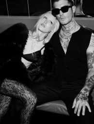Black and white portrait of a blonde woman resting her head on the shoulder of a tattooed man wearing sunglasses.