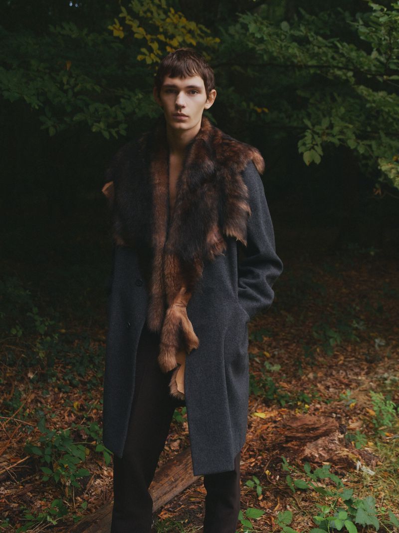 Male fashion model posing in a coat with fur trim in a forest setting, looking directly at the camera with a serious expression.