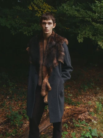 Male fashion model posing in a coat with fur trim in a forest setting, looking directly at the camera with a serious expression.