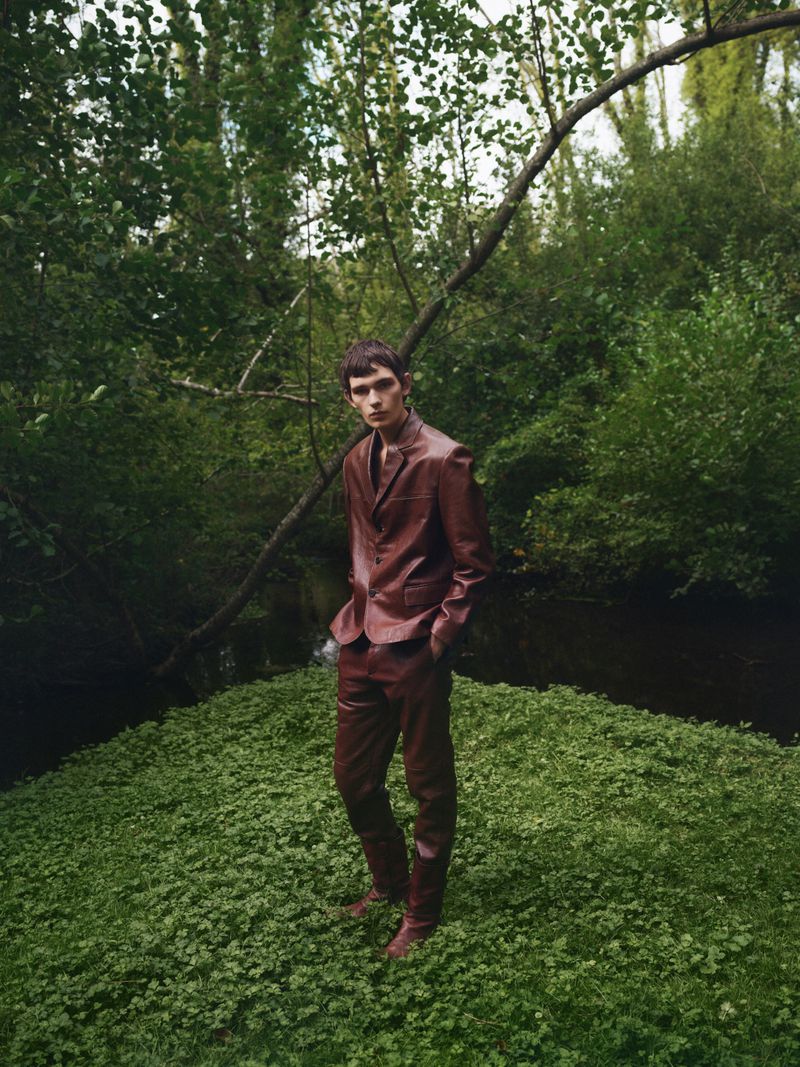 A male model in a red suit stands on green ground surrounded by trees and foliage in a wooded outdoor setting with natural light.