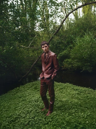 A male model in a red suit stands on green ground surrounded by trees and foliage in a wooded outdoor setting with natural light.