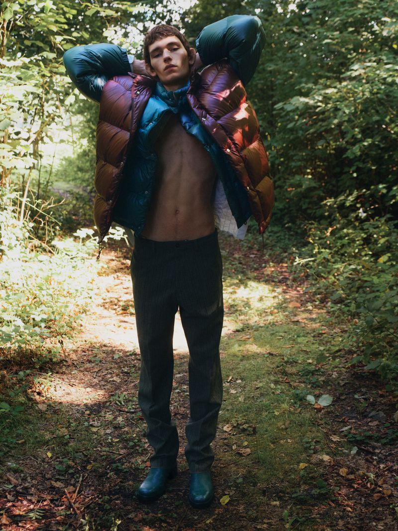 Fashionable adult male poses on a dirt path in a forest wearing an open puffer jacket, pants, and boots in natural light.