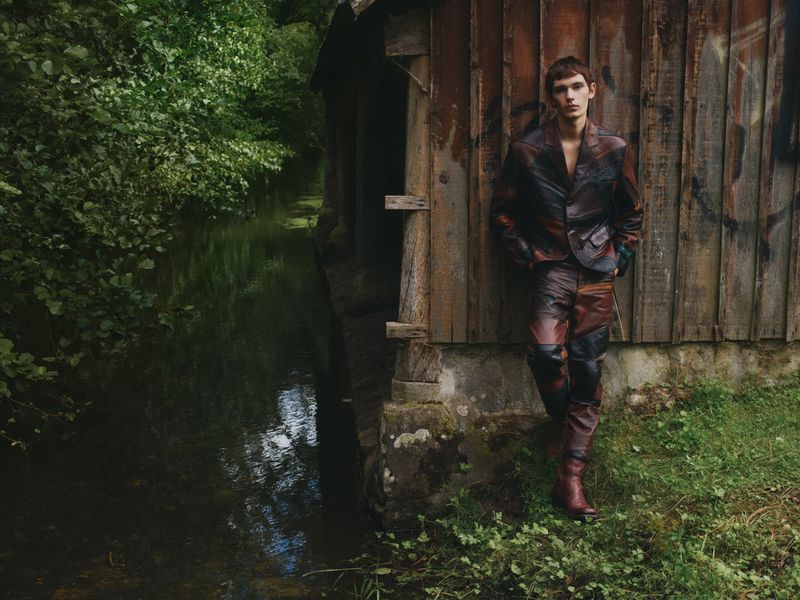 A young male fashion model poses outdoors in a burgundy leather suit, leaning against a rustic wooden shack in a lush, green natural setting.