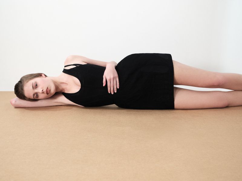 A young woman lies on a tan surface, wearing a black dress with straps, looking toward the viewer in a studio shot with a white backdrop.