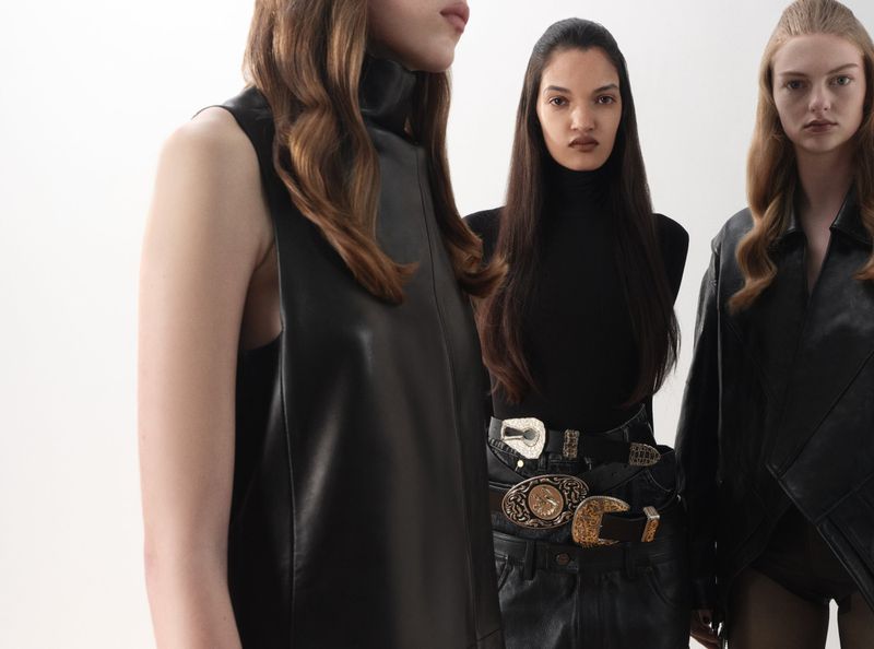 Three female fashion models in black leather outfits pose against a white backdrop. The models wear black turtlenecks and unique belts.