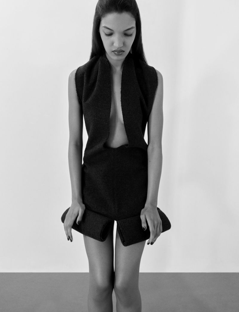 Black-and-white fashion shot of a woman with dark hair, looking down, wearing a black knit top and shorts against a white background.