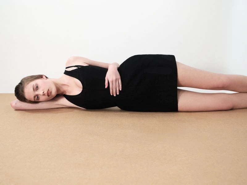 Model Corinne Murray lying on her side in a black sleeveless mini dress against a minimal two-tone studio backdrop.
