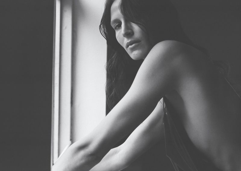 Striking black and white portrait of a woman with long dark hair, posing near a window with natural lighting casting shadows.