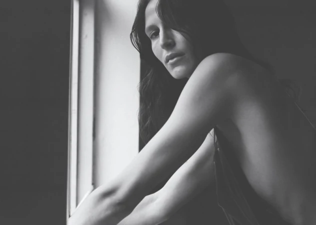 Striking black and white portrait of a woman with long dark hair, posing near a window with natural lighting casting shadows.