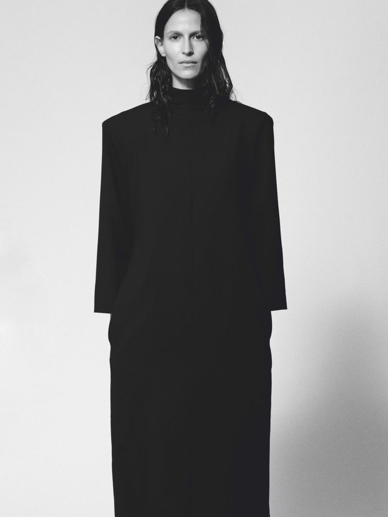 Model Jeanne Cadieu wearing a structured black high-neck dress with padded shoulders against a pale studio backdrop.