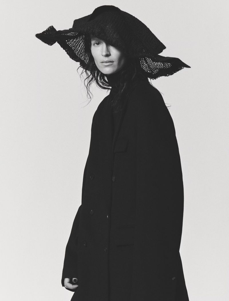 Woman in a black coat and a large, wide-brimmed hat, set against a clean, bright backdrop in a fashion studio environment.