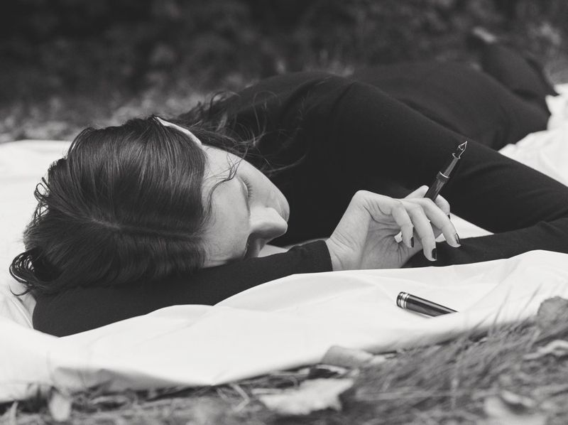 Black and white image of an adult female lying on a white sheet in an outdoor setting, holding a fountain pen, with a contemplative expression.
