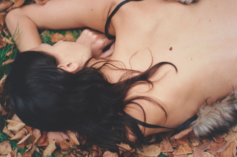 Woman lies on her back outdoors in a field of autumn leaves and green grass, wearing a black top with fur accents on her shoulders.