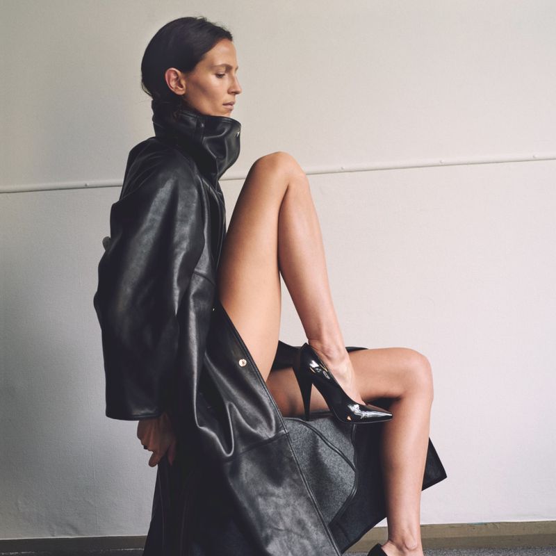 A woman poses indoors wearing a black leather coat and black patent-leather heels. Her leg is raised for a fashion or beauty shoot.
