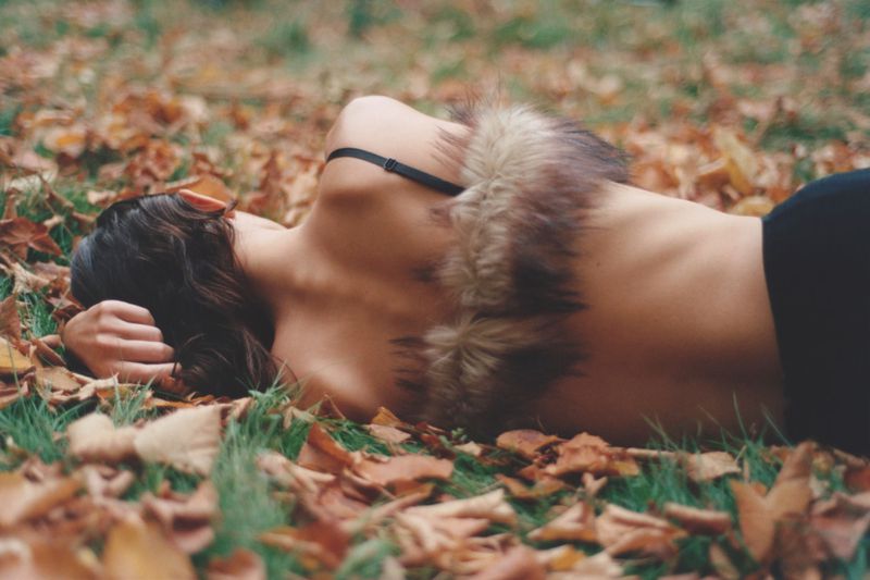 Woman lying on a bed of autumn leaves in a park, wearing a fur collar top. Low-angle close-up shot in natural light.