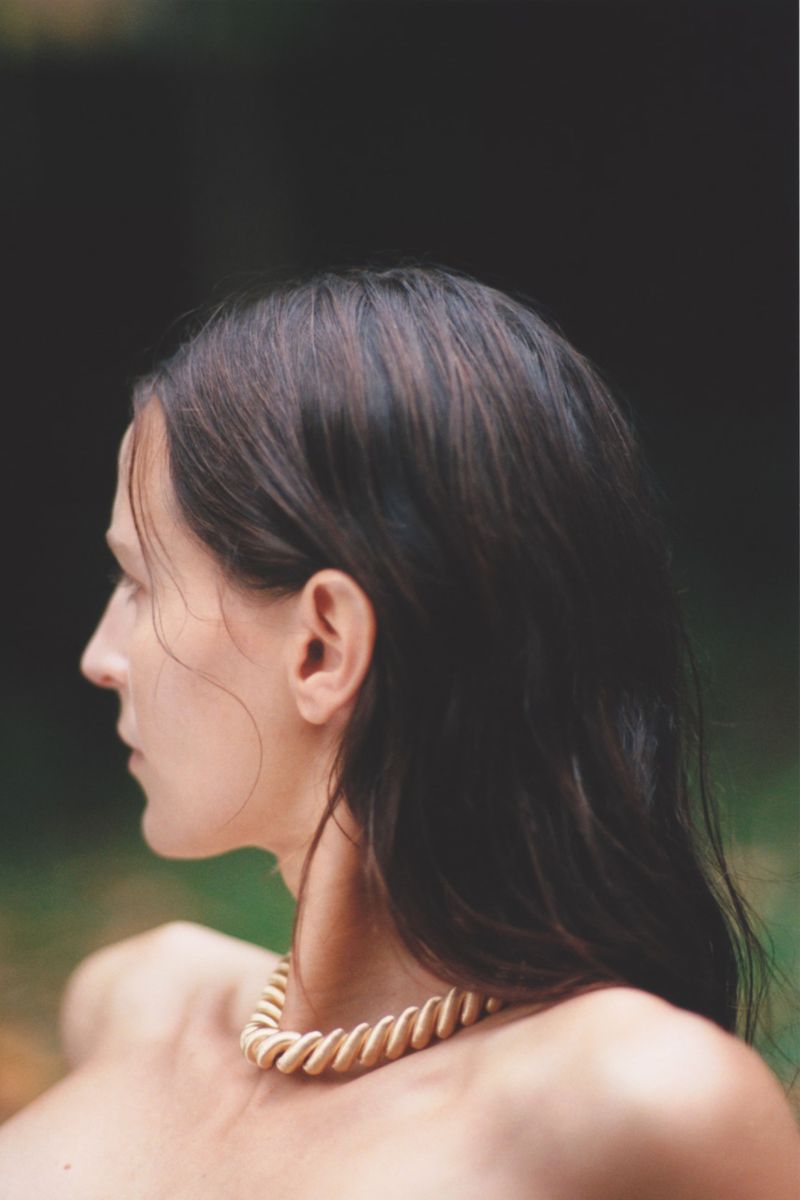 Close-up side profile portrait of a woman with dark hair wearing a gold necklace outdoors against a dark green leafy background.