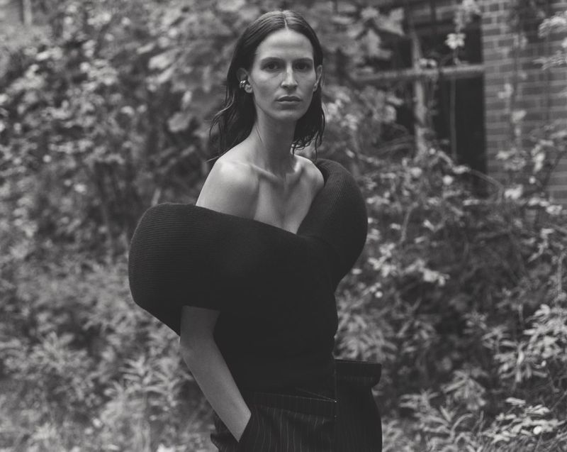Monochrome portrait of a woman in a sculptural black top standing outdoors, with a dense green foliage background, capturing a fashion editorial aesthetic.