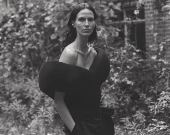 Monochrome portrait of a woman in a sculptural black top standing outdoors, with a dense green foliage background, capturing a fashion editorial aesthetic.