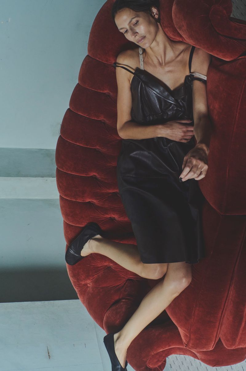 Woman in black dress lying on red velvet couch in indoor studio setting. Stylish fashion photography with unique set design.