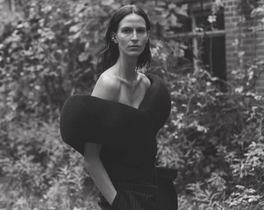 Model Jeanne Cadieu in a sculptural off-the-shoulder black top and pinstripe trousers standing outdoors amid lush foliage.