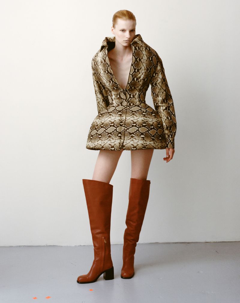 A woman in a snake-print dress and brown boots stands against a white wall. The fashion shot showcases colors and textures.
