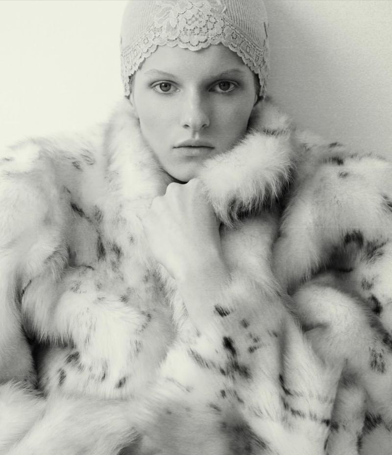 Close-up studio shot of a fair-skinned adult woman wearing a lacy cap and a spotted white fur coat against a bright white backdrop.