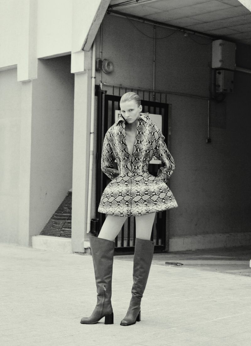 Monochrome fashion shot of an adult woman in a snake-print outfit and knee-high boots standing in front of a garage-like building.