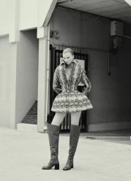 Model in snakeskin print peplum jacket and knee-high boots standing outside an industrial garage entrance.