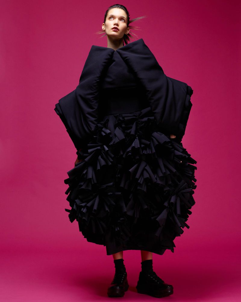 Fashion photo: Woman in oversized avant-garde black dress poses in front of pink backdrop in a studio with creative lighting.